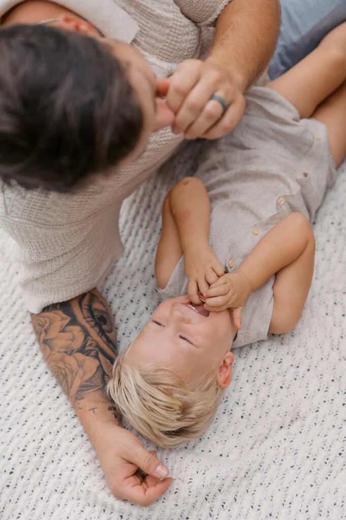 Family session in Kinston, NC, view is from above as dad and toddler boy lay on a blanket, dad tickles toddler boy, toddler boy laughs and had hands in his mouth