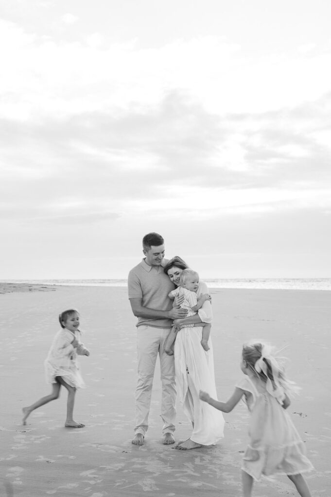 Black and white image of family of five on Emerald Isle Beach at sunrise, dad and mom are snuggled up holding baby as two little girls run around them. 
