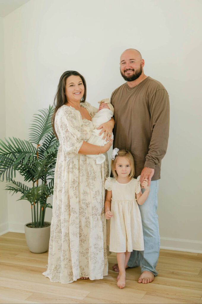 Family of four standing, mom holding baby girl, toddler girl standing in front of dad holding his hand