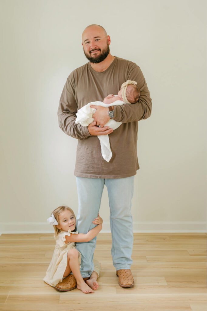Dad standing with toddler girl wrapped around his leg and holding newborn baby girl