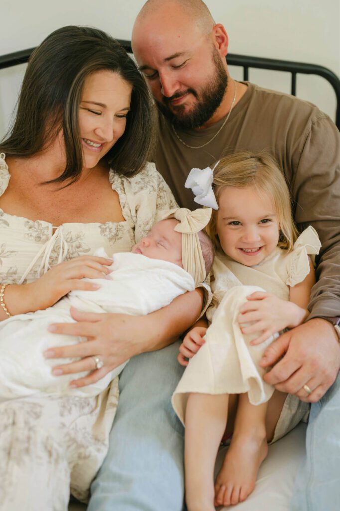 Family of four snuggled up on bed, mom holding baby girl, toddler girl sitting in dads lap smiling
